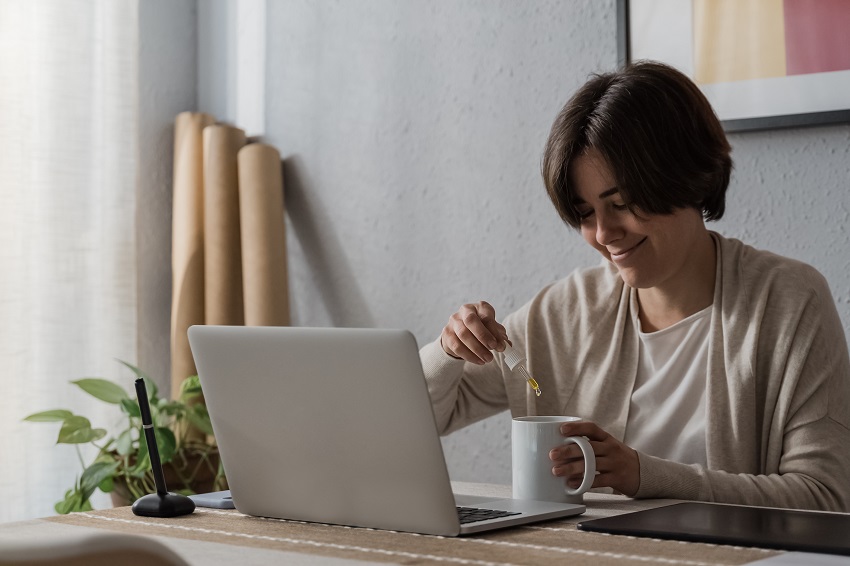 Cbd hemp - Woman taking cannabidiol oil in tea cup while working at home office - Focus on dropper cannabidiol oil in tea cup